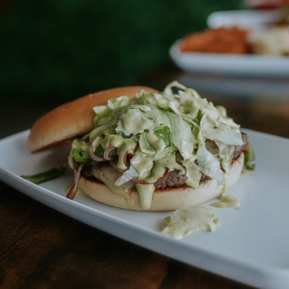 Close-up of a cheeseburger topped with lettuce, onions, vegetables, and creamy sauce on a white plate.
