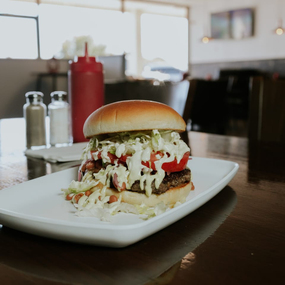 Juicy burger topped with lettuce, tomato, creamy sauce, and onions on a white plate in a sunlit restaurant.