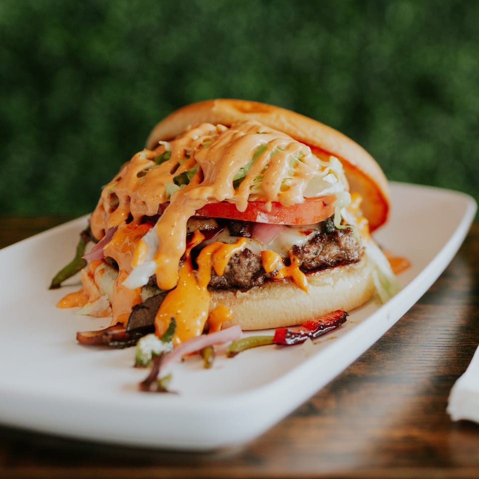 Loaded cheeseburger topped with lettuce, tomato, onions, pickles, and creamy orange sauce on a white plate.