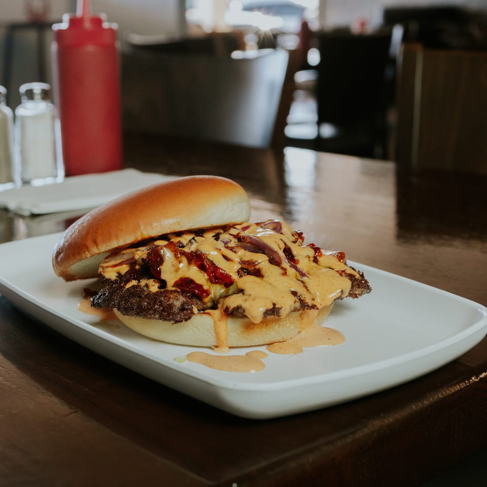Loaded cheeseburger topped with grilled onions, bacon, and creamy sauce on a white plate at a restaurant table.